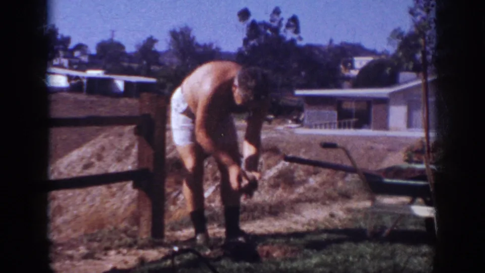 The man bends over the bare ground in a suburban Pennsylvania backyard, with a wheelbarrow and houses behind him.