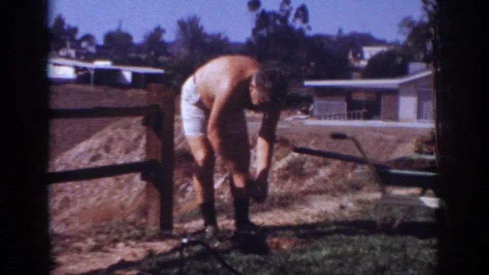 In the 1962 home-movie scene, the man reaches down toward the yard soil while a wheelbarrow sits behind him.