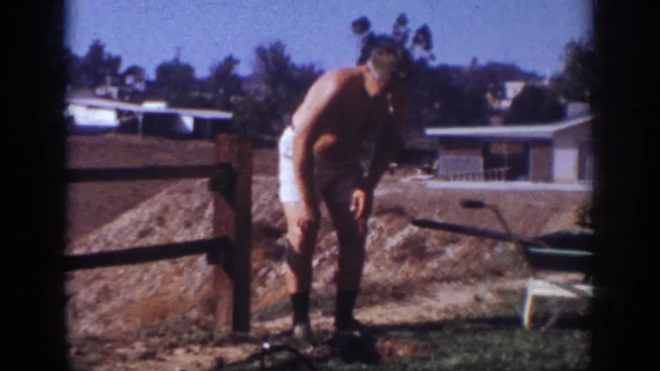 The shirtless man leans forward in the Blainsport backyard, framed by a wheelbarrow, fence, and mid-century houses.