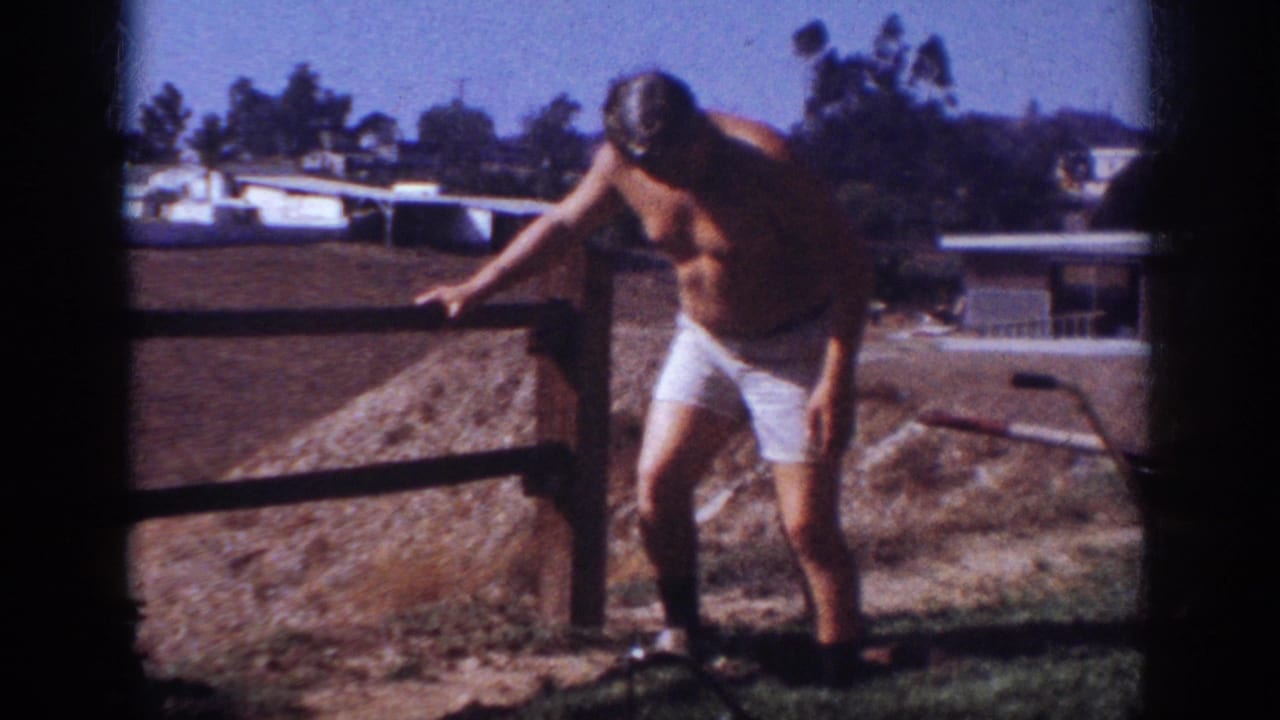 In Blainsport, Pennsylvania, 1962, a shirtless man in white shorts stands near a low wooden fence with dirt and houses behind him.