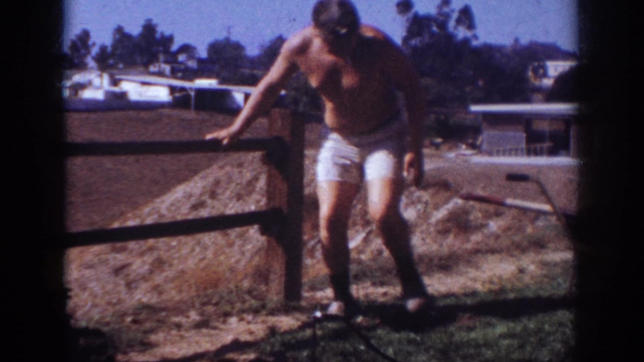 The man steps along the fence line in the 1962 Blainsport home-movie scene, with dry ground, rooftops, and trees behind him.