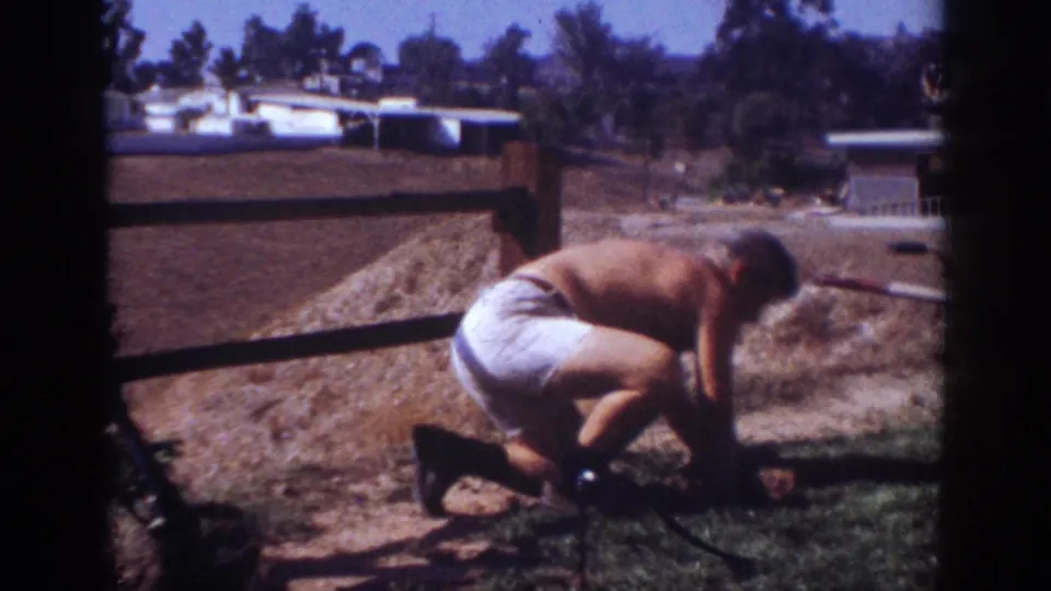 At the end of the 1962 Blainsport clip, the man is crouched in the yard, working close to the dirt beside the fence.