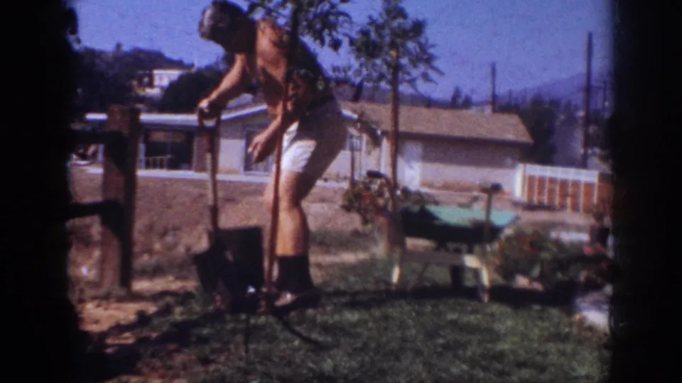 The man steadies the shovel near the young tree in the Pennsylvania backyard, with a wheelbarrow, plants, house, fence, and hills in view.