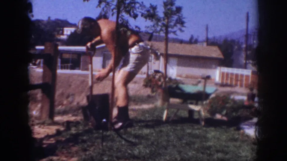 A shirtless man digs at the base of a young tree in the sunny Pennsylvania backyard, framed by soft 8mm film edges.