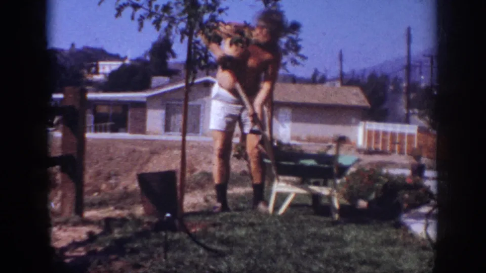 A man in white shorts works with a shovel beside a young tree in a sunny Blainsport backyard, with a ranch-style house behind him.