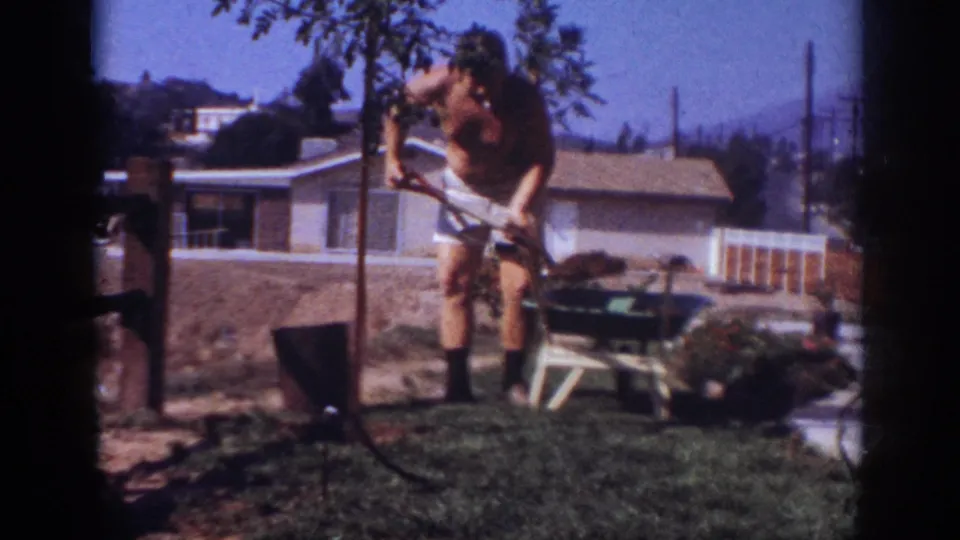 The home-movie frame shows the man digging at the base of a young tree in a suburban Pennsylvania yard in 1962.