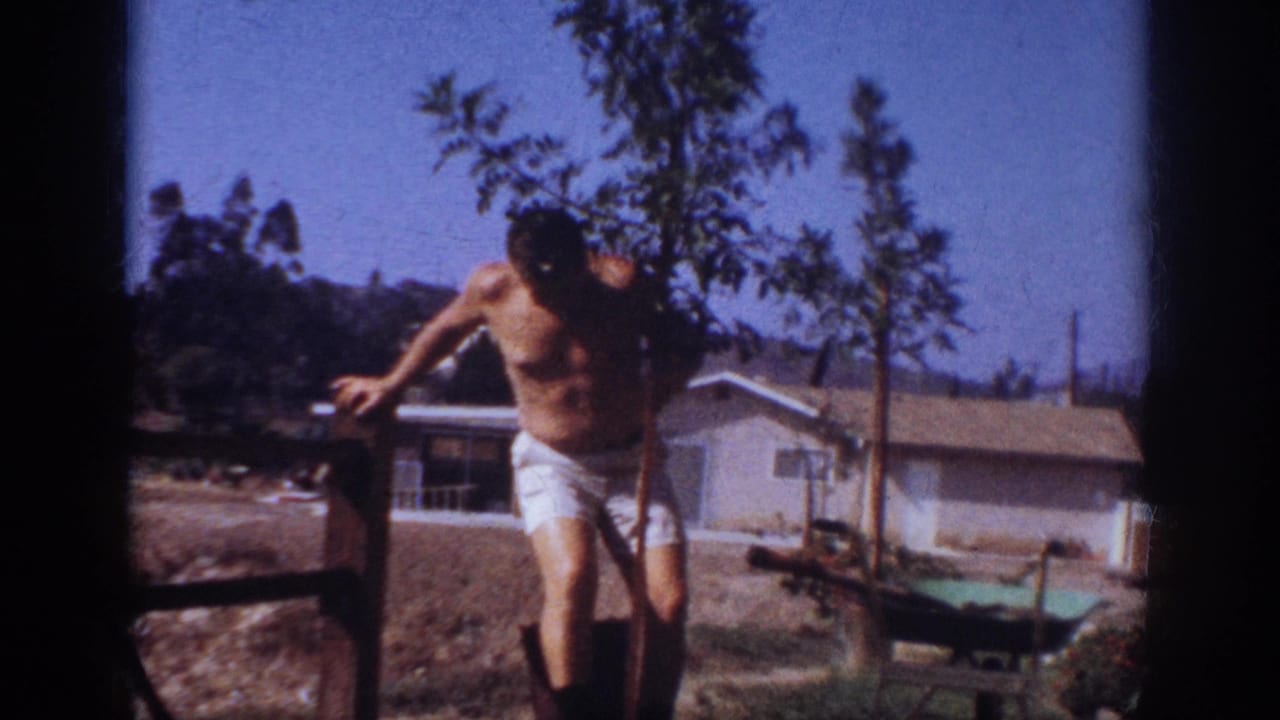 The man works beside the young tree in a 1962 suburban Pennsylvania backyard, with a low house and wheelbarrow behind him.