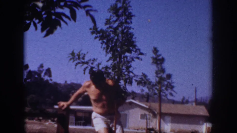 A shirtless man bends near the sapling during yard work in Blainsport, Pennsylvania, in 1962, framed by home-movie film grain.