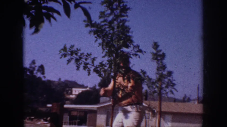 1962 Blainsport home-movie frame showing a shirtless man near a sapling, with rooftops, hillside homes, and blue sky behind him.