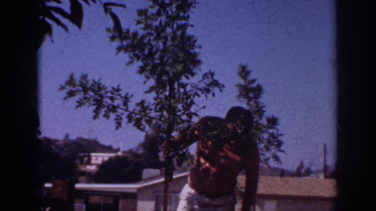 1962 Blainsport frame of a man leaning toward a young tree in a backyard, with a low roofline and distant hillside visible.