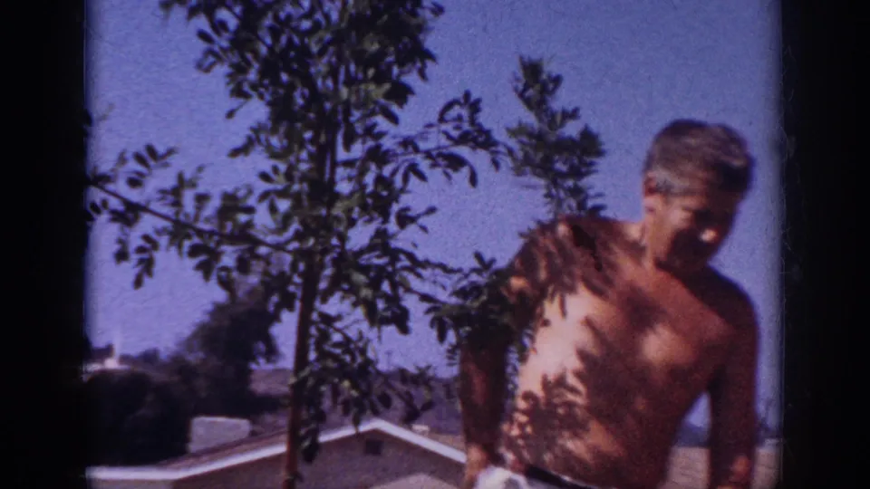 The man turns beside the young tree near the end of the Blainsport backyard clip, with rooftops, hills, and clear sky in view.