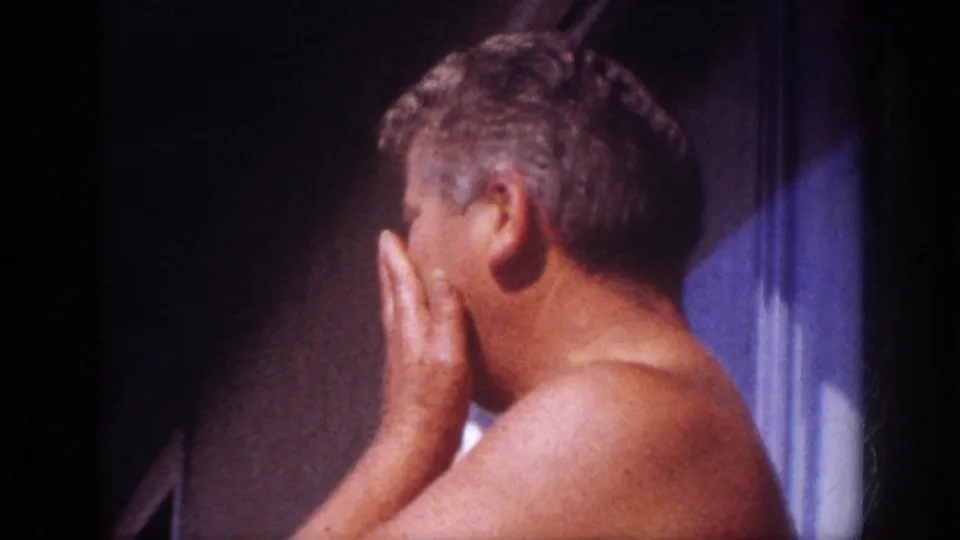 Blainsport 1962 frame showing a shirtless adult man in profile with his hand near his face beside a shaded exterior wall.