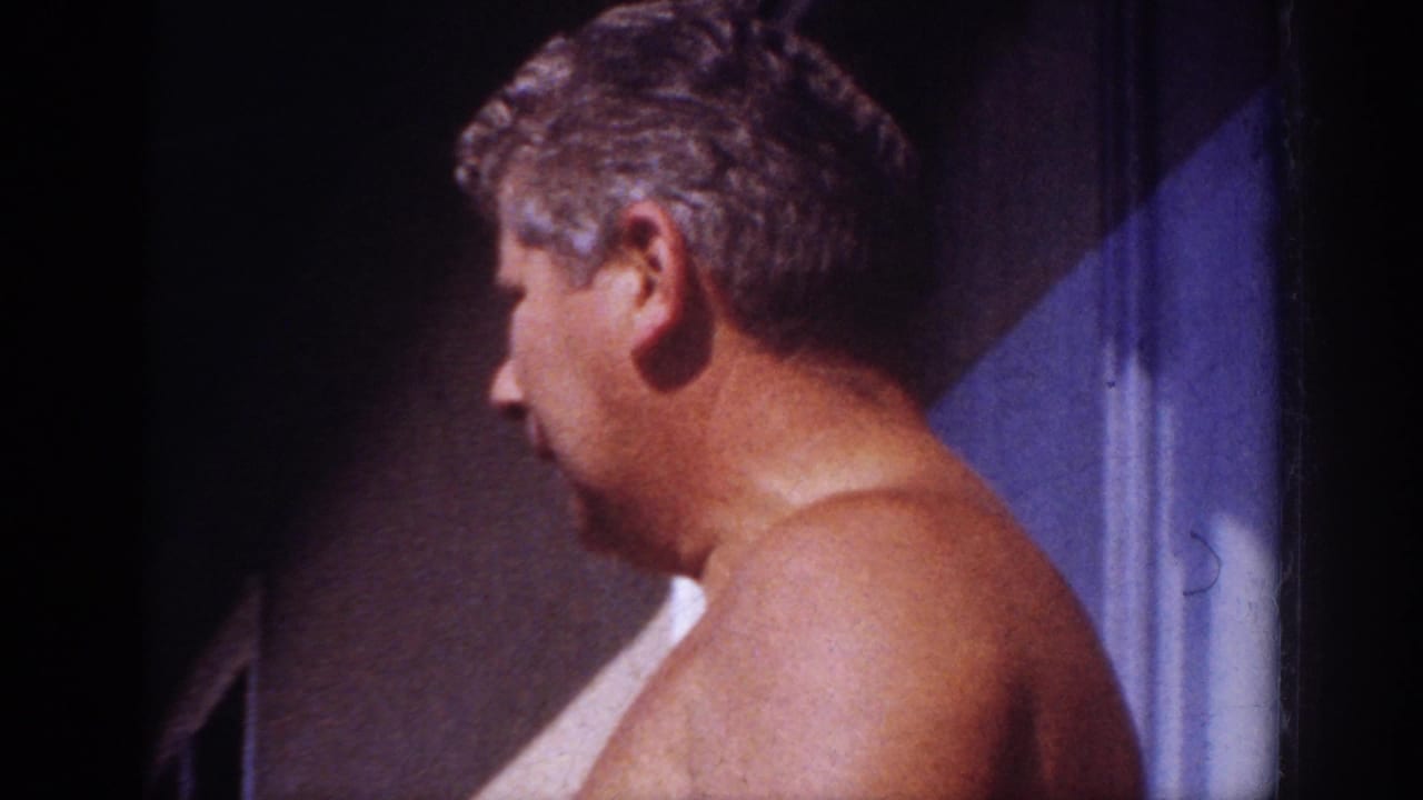 In 1962 Blainsport home-movie footage, a shirtless adult man stands in profile near a doorway or wall at the edge of a yard.