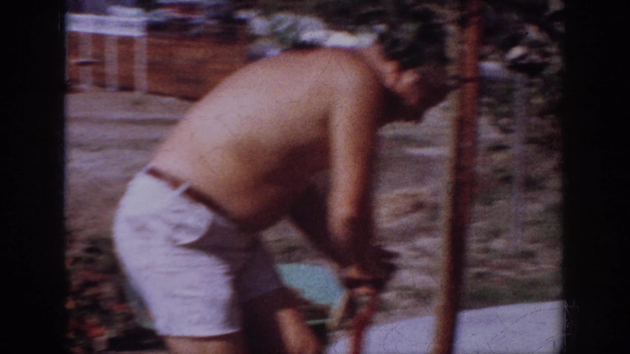 The man bends over in a Blainsport backyard, working near a young tree or support stake with houses and fencing behind him.