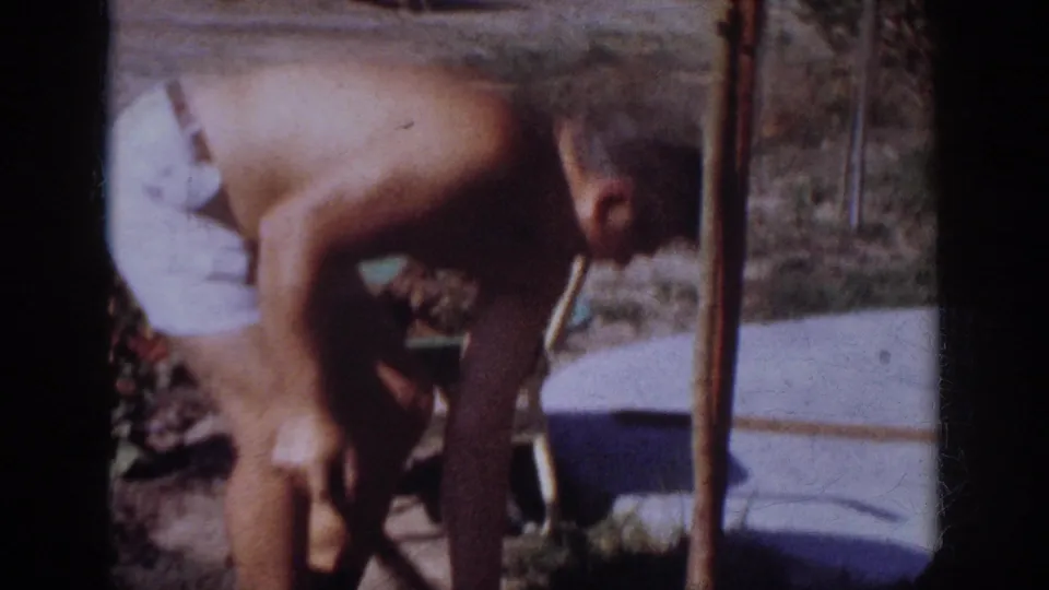 A closer 8mm frame shows the man bent low beside a vertical stake, with grass, soil, and a low wall behind him.