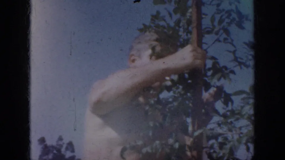 1962 Blainsport home-movie frame showing a shirtless man outdoors beside foliage and a vertical wooden element.