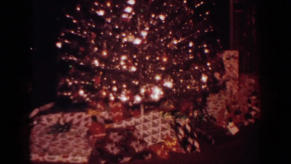 Home-movie frame from the 1962 clip showing a lit Christmas tree and wrapped presents in a dark interior.