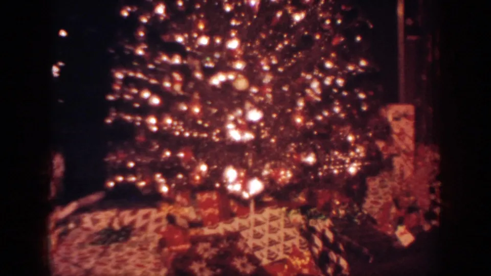 1962 Blainsport home-movie frame showing a glowing Christmas tree, ornaments, and multiple wrapped gifts beneath it.