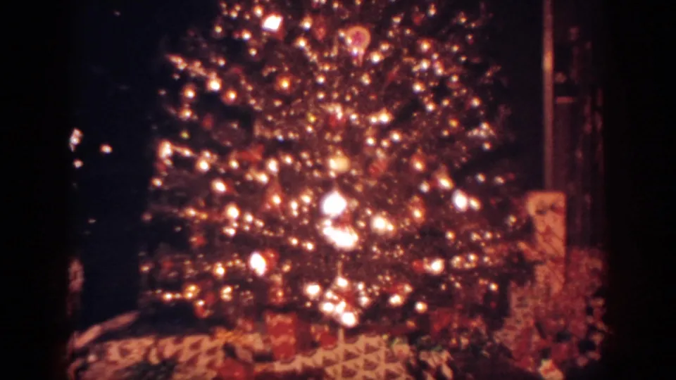 Home-movie frame showing the illuminated branches of a Christmas tree with wrapped presents visible along the bottom edge.