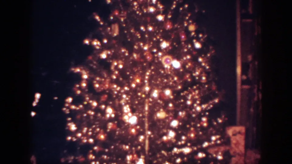 Vertical view of the decorated Christmas tree, showing dense lights, ornaments, and dark room edges in 8mm film texture.