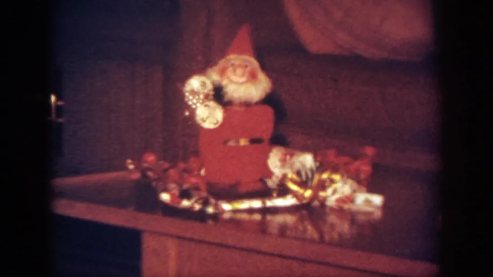 Close 8mm frame of a red Santa figurine with a white beard beside tinsel and small holiday items.