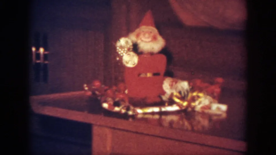 Tight 1962 Blainsport frame of a small Santa figurine on a table, with tinsel and holiday objects around it.