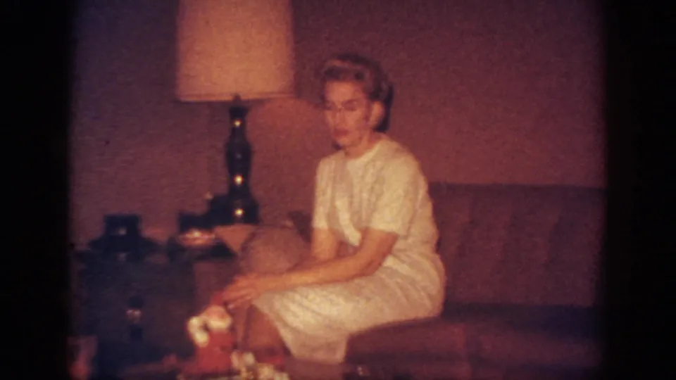 1962 Blainsport home-movie frame of a blonde woman in a white dress seated on a sofa beside a lamp and tabletop Christmas decoration.