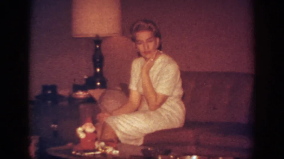 1962 Blainsport home-movie frame showing a blonde woman seated on a sofa, posing near a lamp and small Christmas tabletop decoration.