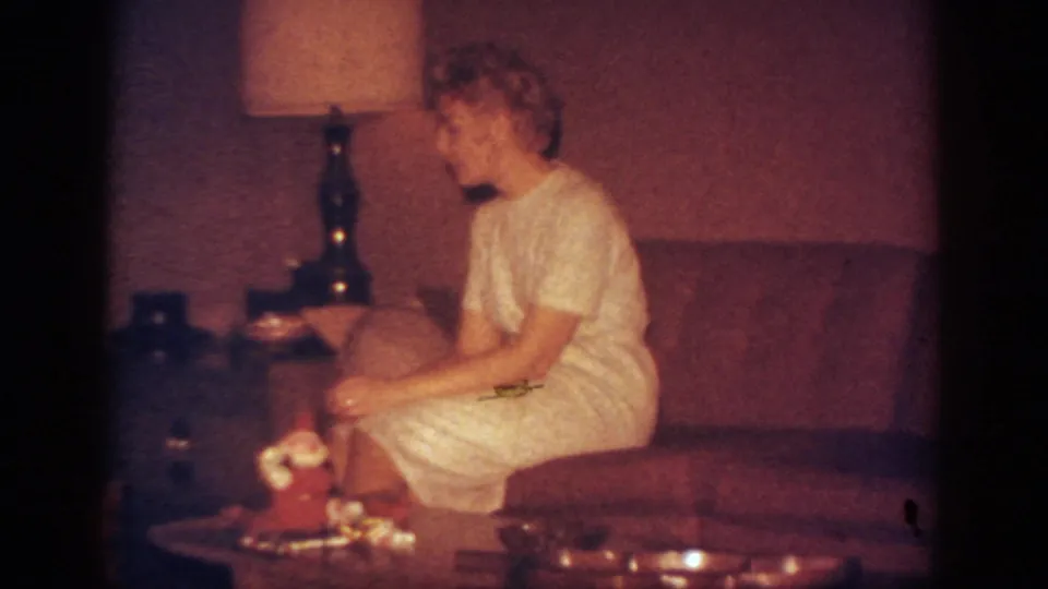 1962 Blainsport home-movie frame of a woman in profile on a sofa, with a lamp and Christmas tabletop decoration in front of her.