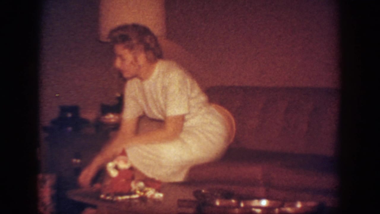 In a 1962 Blainsport home-movie frame, a woman in a light dress leans from a couch toward presents on a coffee table.