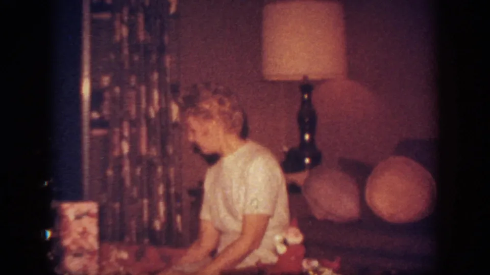 A woman sits beside Christmas gifts in a mid-century living room, with lamp, curtains, couch, and round cushions visible.