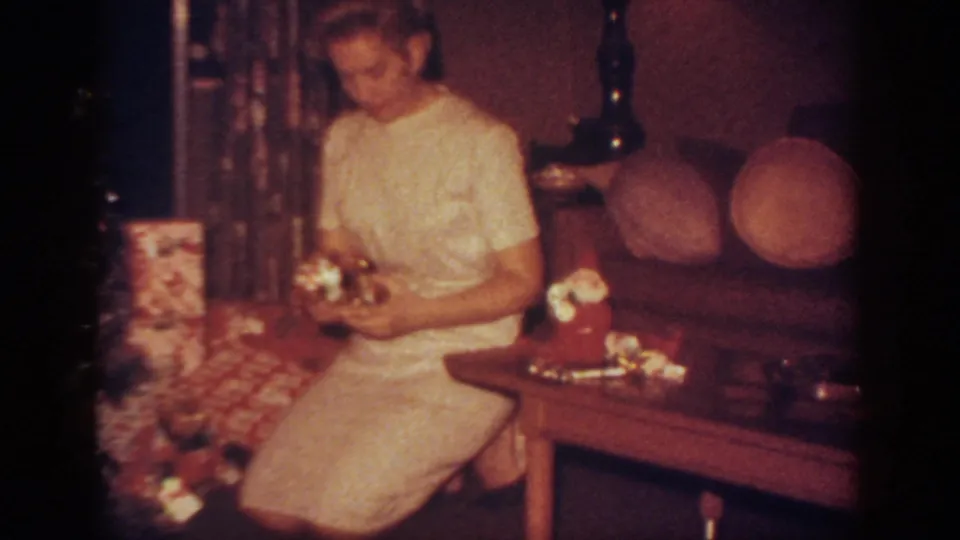 The woman holds Christmas gifts beside the coffee table in the Blainsport living room, with couch cushions and lamp behind her.