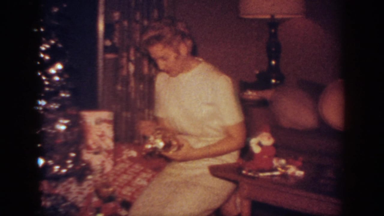 Woman in a white dress sits beside a lit Christmas tree and wrapped gifts in a living room.
