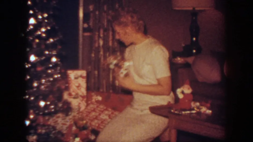 The woman turns toward the Christmas tree in the Blainsport living room, with wrapped gifts, curtains, a lamp, and a side table visible.