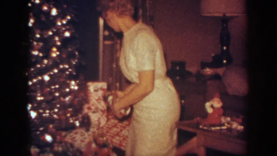 In the 1962 home-movie frame, the woman stands beside the glowing Christmas tree and reaches down toward wrapped gifts in the living room.