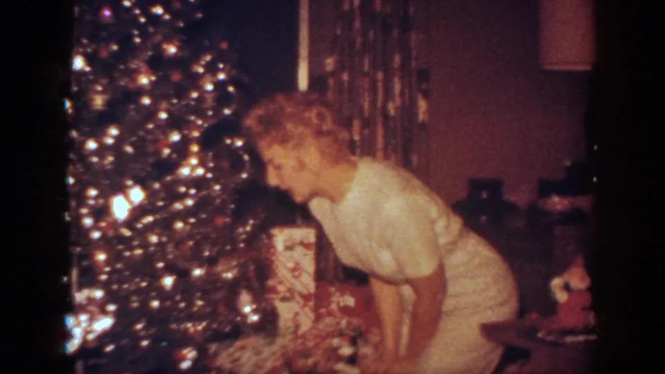 The woman bends near the base of the Christmas tree, with lights, wrapped gifts, curtains, a lamp, and side furniture visible in the Blainsport home.