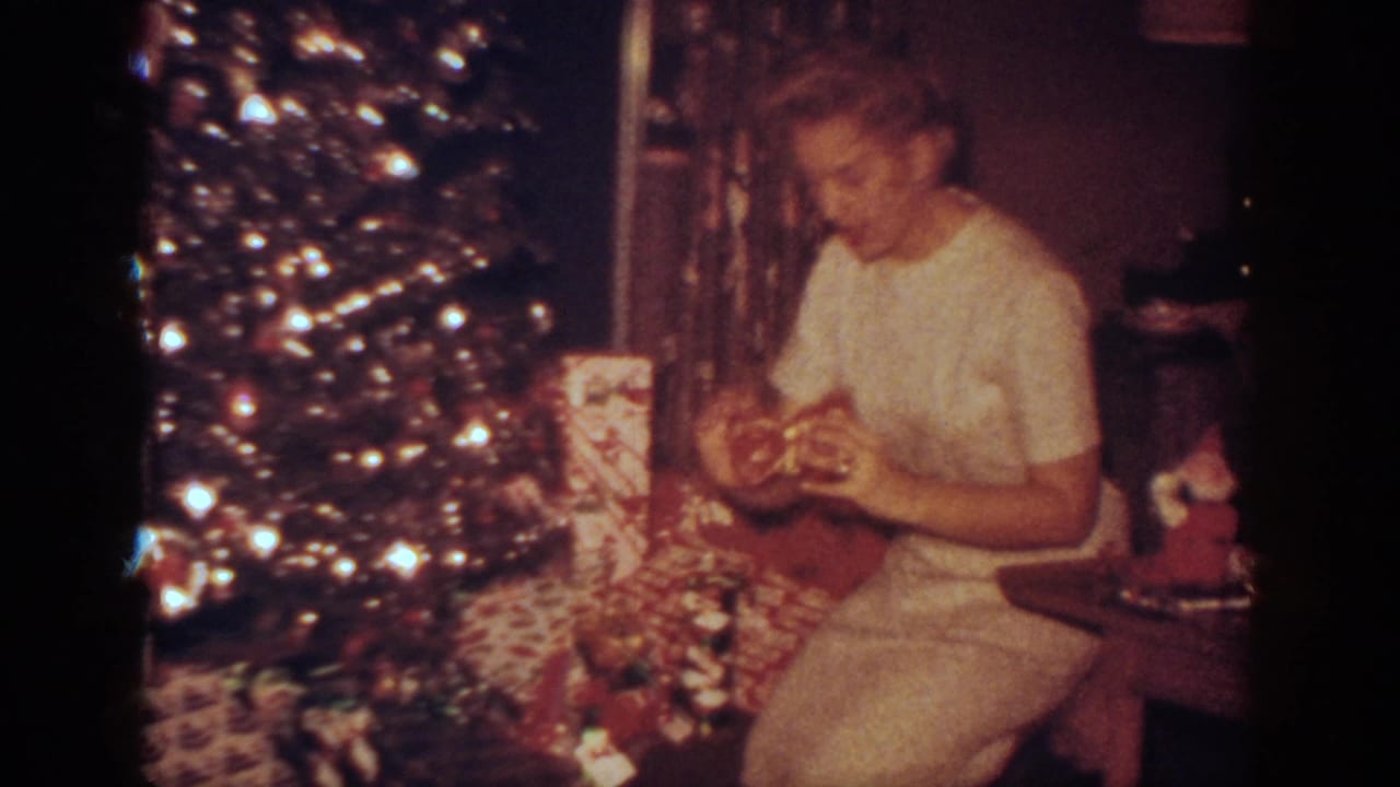 Seated woman holds a wrapped present beside a brightly lit Christmas tree.