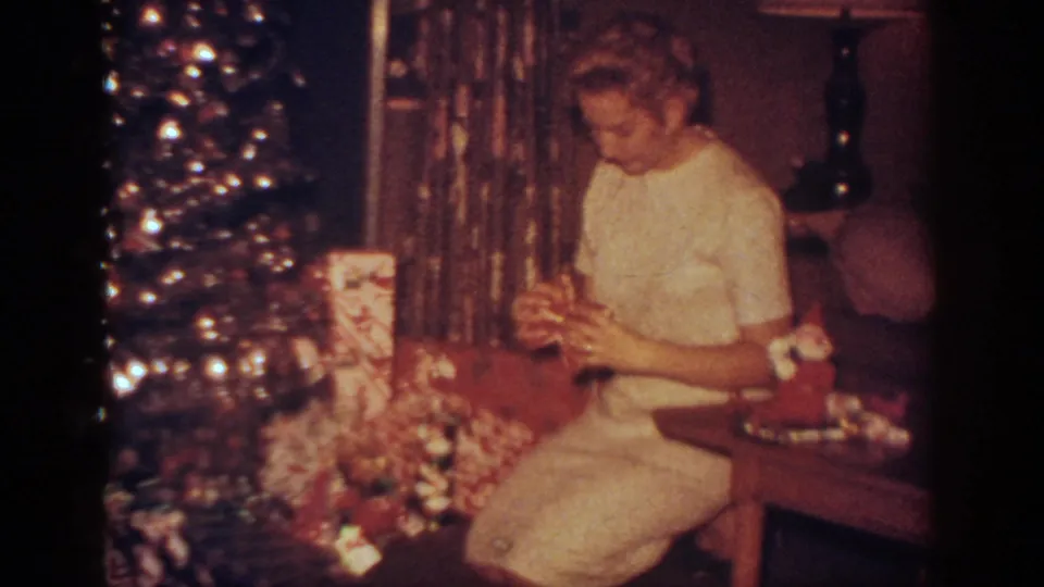 The woman continues examining a wrapped package beside the Christmas tree and gift pile in the Blainsport holiday interior.