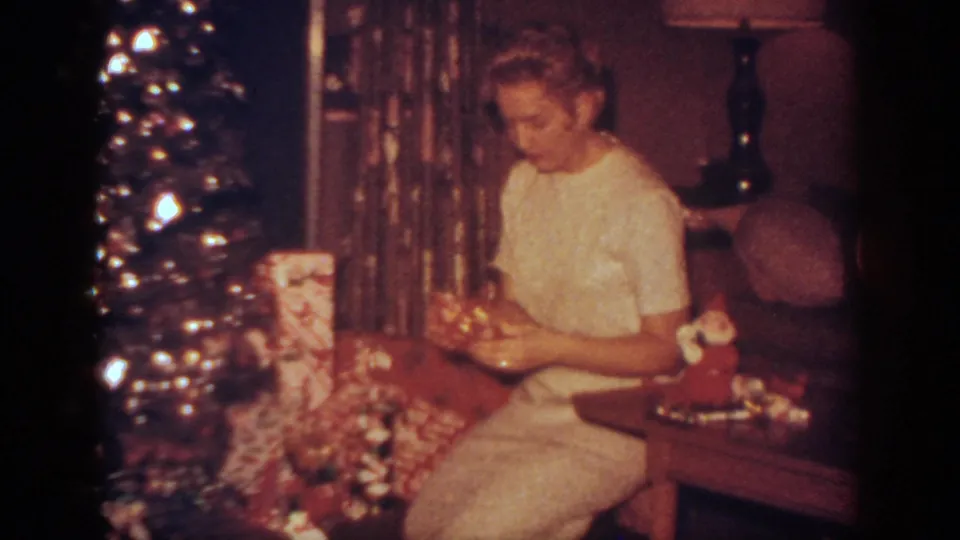 A woman seated in a white dress looks down at a present near the Christmas tree, with wrapped gifts and household furnishings around her.