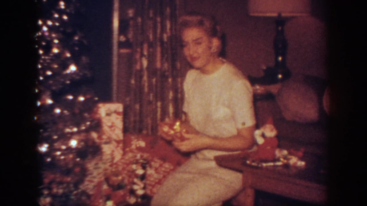 In a 1962 Blainsport home interior, a woman in a white dress sits near a lit Christmas tree with wrapped gifts and decorations.