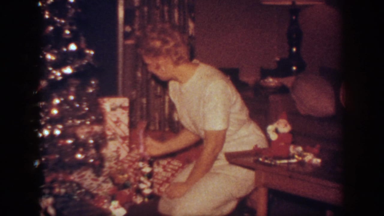In a 1962 Blainsport home interior, a woman in light clothing kneels beside a shimmering Christmas tree and wrapped gifts.