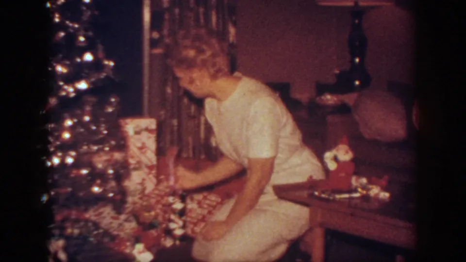 In a 1962 Blainsport home interior, a woman in light clothing kneels beside a shimmering Christmas tree and wrapped gifts.