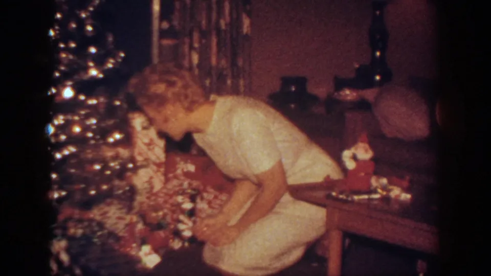 The woman bends toward gifts under the shimmering Christmas tree in the dimly lit Blainsport holiday interior.