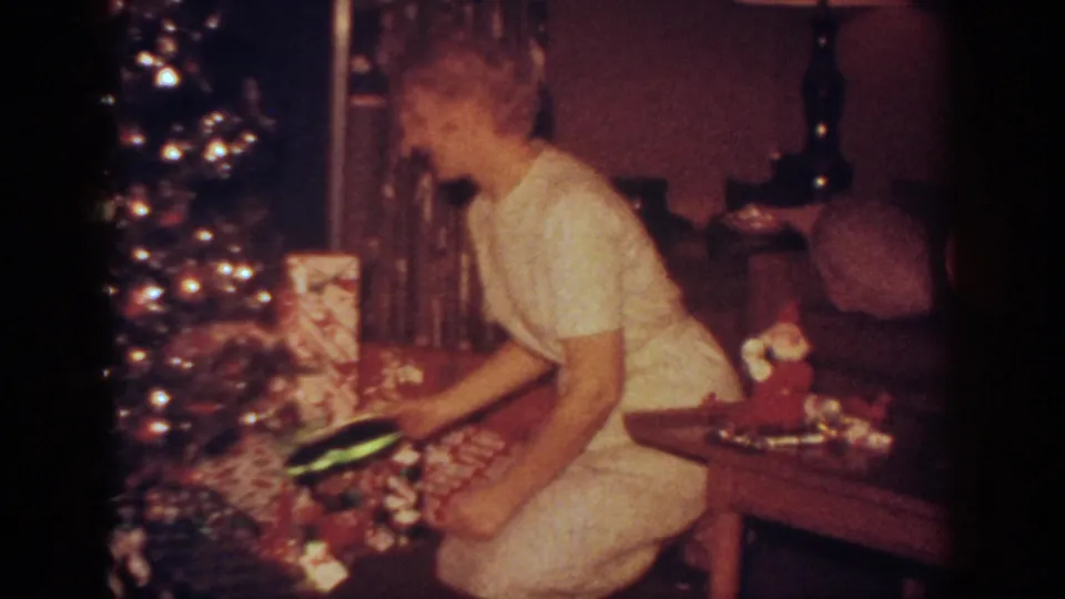 The woman appears to lift or inspect a small present beside the Christmas tree, with a Santa decoration on the nearby table.