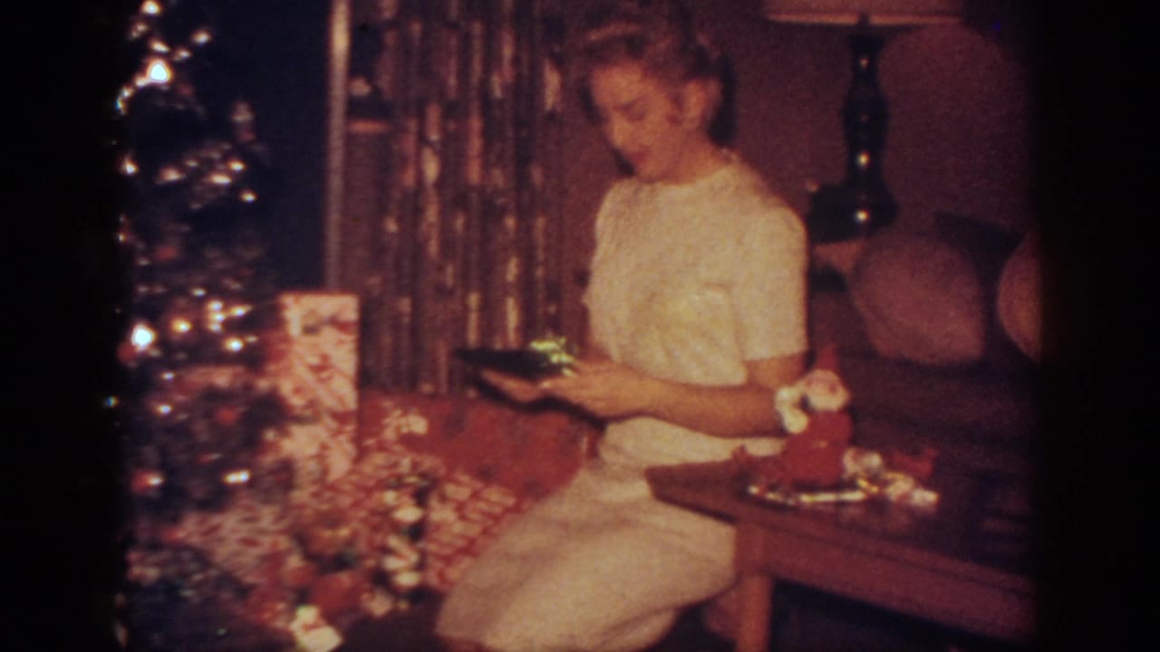 In this 1962 Blainsport, Pennsylvania home movie frame, a woman in a white dress sits by a Christmas tree while holding a gift near wrapped presents.