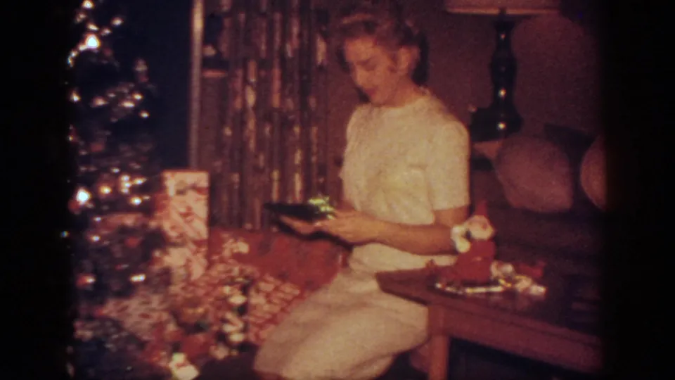 In this 1962 Blainsport, Pennsylvania home movie frame, a woman in a white dress sits by a Christmas tree while holding a gift near wrapped presents.