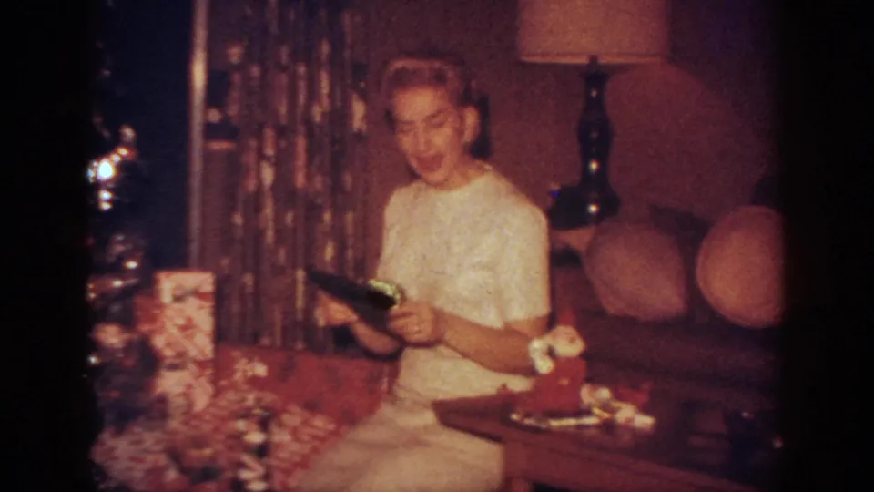 A woman in a white dress appears to smile while holding a small gift beside a decorated Christmas tree in a 1962 Blainsport home interior.