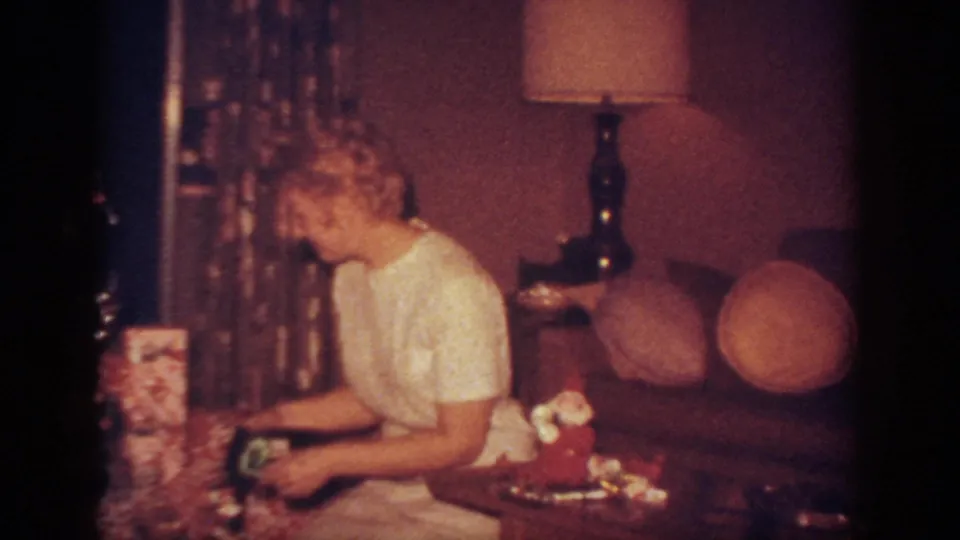 The woman leans down toward the presents near the Christmas tree, with tinsel curtains, a table lamp, and a chair visible in the home interior.