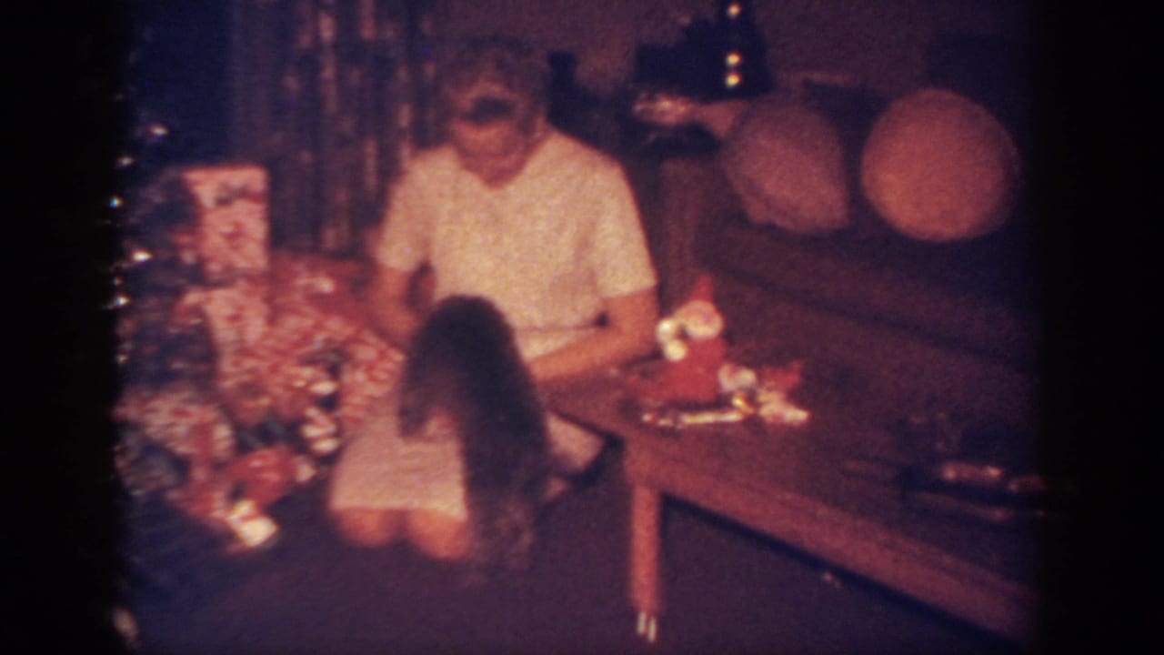 In a 1962 Blainsport, Pennsylvania home-movie frame, a woman kneels near wrapped gifts as a small black puppy faces her.