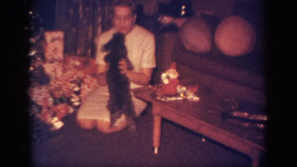 In the holiday living room, the woman lifts the small black puppy while kneeling beside wrapped presents and a coffee table.